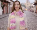 Mujer sonriente luce bufandas de dos agujas modernas en tonos pastel, paseando por una calle empedrada.