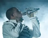 Artysta w białej kurtce z czarnymi zdobieniami śpiewa do mikrofonu. Te rap wspomnienia na zawsze w sercu.