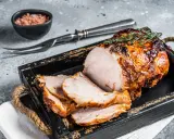 A sliced, roasted pork loin with a golden-brown crust, garnished with thyme, on a rustic wooden tray.