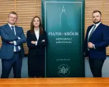 Trzy osoby w garniturach stoją przed zielonym banerem kancelarii adwokackiej Piątek-Krolik.