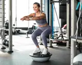 Frau macht Kniebeugen auf einem Bosu-Ball, trägt Sportkleidung und weiße Sneaker. Sie trainiert ohne schuhe auf dem instabilen Untergrund.