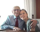 Uśmiechnięty starszy mężczyzna w okularach i młoda kobieta przytulają się. Widać, jak różne generacje ludzi łączą się w ciepłym uścisku.