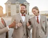 Groomsmen in tan suits with rust ties and bowties toast with beers. A great color suit for wedding celebrations.