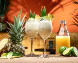 Två krämiga drinkar med ananasjuice, is och lime. En hel och en halv ananas samt en flaska juice står bredvid.