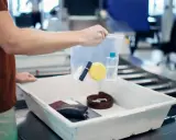 Ręka trzyma przezroczystą torebkę z kosmetykami nad tacą z rzeczami do kontroli bezpieczeństwa na lotnisku.