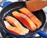 Vier Lachsfilets braten in einer Pfanne. Ein Holzspatel liegt bereit, um zu zeigen, wie bereite ich Lachs zu.