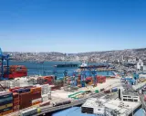 Un vasto porto con gru, container colorati e navi cargo, con una città collinare sullo sfondo. Sembra il porto più grande del mondo.