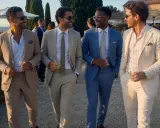 Four stylish men in suits and chinos walk together at a wedding, holding drinks and smiling.