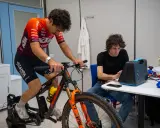 Ciclista su rulli monitorato per l'acido lattico, con un tecnico che osserva i dati su un dispositivo.