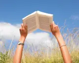 Mani che sorreggono un libro aperto contro il cielo azzurro, tra fili d'erba alta. Un momento di pace e apprendimento, magari con libri sulla salute mentale.