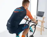 Mężczyzna w stroju kolarskim pedałuje na rowerze stacjonarnym, śledząc trasę na ekranie laptopa z aplikacją Zwift.