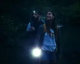 Para idzie przez las nocą, oświetlając drogę latarką. Mężczyzna ma czołówkę, a kobieta trzyma mocny **sos sygnal**.