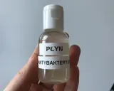 Dłoń trzyma małą, przezroczystą butelkę z białym zamknięciem, zawierającą płyn.