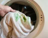 Hand hält Handtuch mit grünen, eingewaschenen Flecken vor einer Waschmaschine.