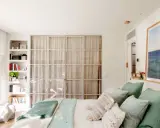 Estantería blanca aprovecha hueco entre cama y pared, con libros y plantas. Puertas de cristal con cortinas separan el espacio.