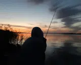 Zachód słońca nad jeziorem, wędkarz czeka na branie. Zastanawia się, jaka będzie karta wędkarska cena.