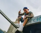 Uśmiechnięty raper z tatuażami, w czapce i kolorowej koszulce, siedzi na dachu, promując swój mixtape "sos ciuchy i borciuchy".
