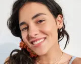 Primo piano di una giovane donna con un sorriso radioso, la sua fronte lucida riflette la luce. Indossa orecchini a cerchio dorati e una collana sottile.