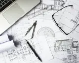 Na stole architekta leżą plany budynku, laptop, okulary i cyrkiel. To miejsce pracy nad projektem i jego kosztorysem.
