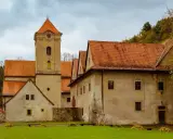 Czerwony klasztor w Pieninach, z charakterystyczną wieżą zegarową i pomarańczowymi dachami, otoczony zielonym trawnikiem i drzewami.