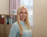 Uśmiechnięta blondynka w jasnej koszuli, za nią regały z książkami.