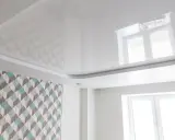 Biały, błyszczący sufit napinany z subtelnymi wzorami geometrycznymi, odbijający światło okna.
