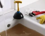 Un évier bouché après siphon, avec de l'eau sale stagnante. Un déboucheur, une clé à molette rouge et des gants jaunes sont prêts à l'emploi.
