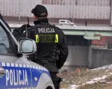 Policjant w kamizelce z napisem "POLICJA", zgodnej z rozporządzeniem w sprawie umundurowania policjantów, stoi obok radiowozu.