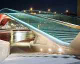 Le pont de la Constitution, illuminé la nuit, avec ses escaliers de verre et ses arches rouges, surplombe un canal aux reflets lumineux.