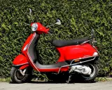 Scooter rojo brillante aparcado frente a un seto verde. ¡Listo para salir con mi permiso AM!