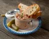 Un pot de rillettes de porc, garni d'une brindille de romarin, est servi avec des cornichons et des tranches de pain croustillant sur une assiette décorée.