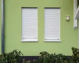 Dwa okna z białymi roletami zewnętrznymi na zielonej ścianie domu. Różne rodzaje rolet zewnętrznych chronią przed słońcem i zapewniają prywatność.