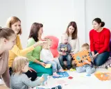 Grupa dzieci i dorosłych uczestniczy w zajęciach, które przypominają terapię pedagogiczną. Dzieci bawią się klockami i zabawkami.