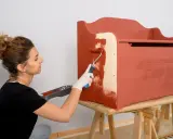 Mujer con guantes blancos pinta con rodillo una caja de madera de color terracota.