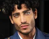 Primo piano di un uomo con splendidi capelli ricci scuri e occhi azzurri penetranti, indossa una giacca blu.
