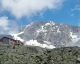 Rifugio Gherardi, un edificio con finestre rosse, si erge su un pendio roccioso sotto un imponente massiccio montuoso innevato.