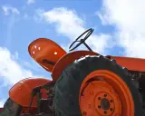Close-up of an orange tractor's wheel and seat against a blue sky with clouds. This vintage tractor, possibly a different size Kubota tractor, awaits its next task.