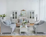 Salotto in stile shabby chic con divani grigi, poltrone, tavolino bianco, specchio dorato e vetrine bianche.
