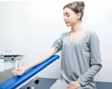 Młoda kobieta czeka na pobranie krwi. Jej ręka spoczywa na niebieskim podłokietniku. W tle widać stojak z probówkami.