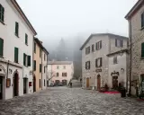 Piazza di San Leo avvolta nella nebbia, con edifici in pietra e intonaco, un bar e una farmacia.