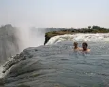 Zwei Personen schwimmen im Devils Pool am Rande der Victoriafälle, mit Gischt und üppiger Vegetation im Hintergrund.