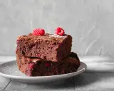 Dwa pyszne brownie z malinami, posypane cukrem pudrem, ułożone na szarym talerzyku.