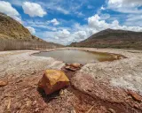 Un estanque natural en Les Salinetes de Novelda, rodeado de rocas y vegetación seca, bajo un cielo azul con nubes blancas.