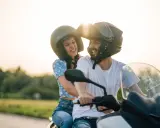 Pareja sonriente en moto, ella con blusa floral y él con camiseta blanca. Listos para disfrutar de la carretera con su carnet A2.