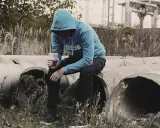 Chłopak w niebieskiej bluzie siedzi na betonowych rurach, trzymając kubek z napojem. W jego oczach widać smutek, jakby czuł, że dda nie umiem kochać.