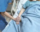Pielęgniarka trzyma za rękę pacjentkę leżącą w łóżku szpitalnym pod niebieskim kocem.