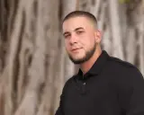 Mężczyzna z buzz cut z zakolami i brodą, w czarnej koszulce polo, na tle drzew.