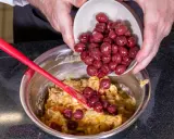 Hände geben Kirschen in eine Schüssel mit Teig für ein einfaches Kirschenmichel Rezept. Ein roter Teigschaber liegt daneben.