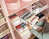 Frau sortiert Kleidung in einem rosafarbenen Schrank. Die Fächer sind ordentlich mit gefalteten Kleidungsstücken gefüllt, die nach Farben sortiert sind, um eine harmonische Reihenfolge zu schaffen.