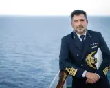 Il comandante MSC Splendida, con la sua uniforme blu scuro e il cappello dorato, posa con il mare calmo sullo sfondo.
